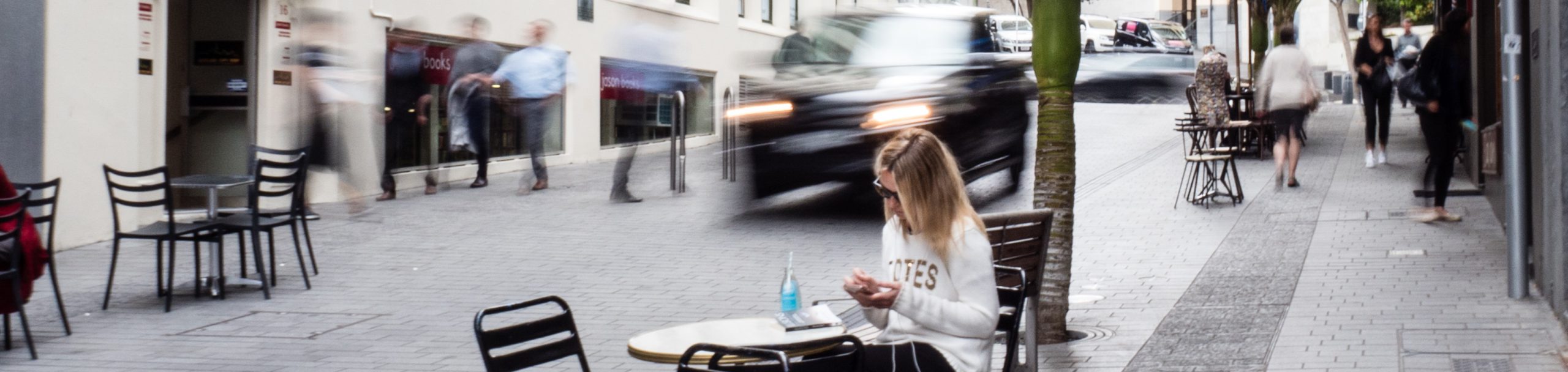 A woman sits at a cafe table on a shared street while a car passes behind her